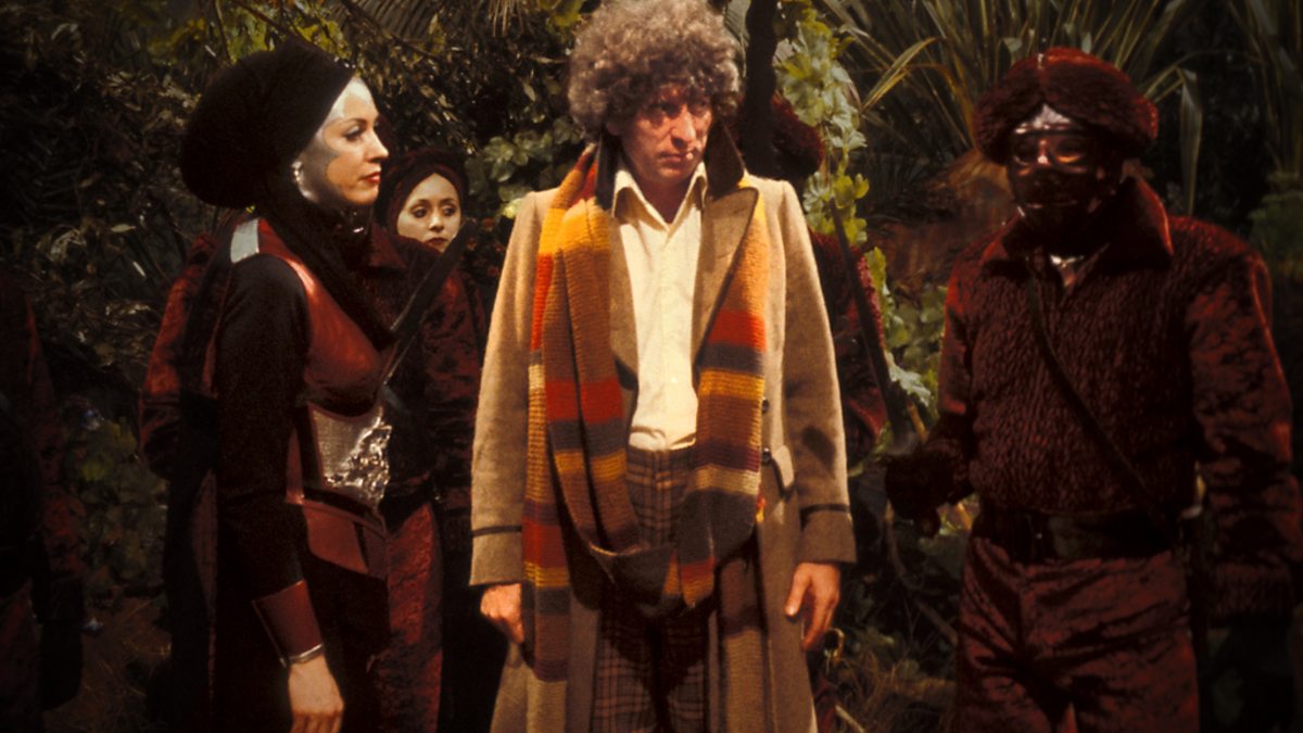Screencap from The Creature from the Pit: The Doctor is flanked by red-armored guards and Adrasta, a woman in a head wrap and silver makeup that outlines her face.