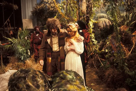In an jungle, the Doctor has his arm around Romana as they look down on two wolfweeds: round dark green plants about two feet in diameter. The edge of the set is visible to the side.