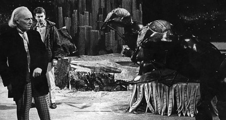 Screencap from The Web Planet: on barren terrain, the First Doctor (with Ian behind him) look on at several Zarbi. The Zarbi are rather obviously people in long horizontol ant costumes, with human legs sticking out from their middles.