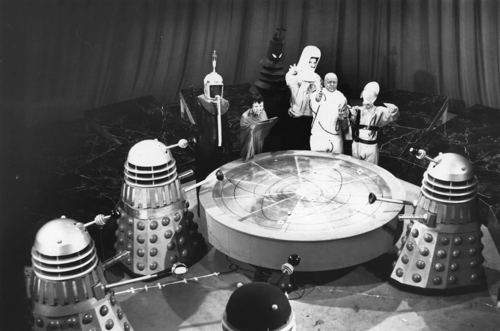 Screencap from Mission to the Unknown (episode one of Nightmare on Planet Kembel): a wide open conference room shows 6 characters and 3 Daleks (as well as one black Dalek) around a round table with some sort of star map on it, viewed from above. The alien characters are (from left to right): a person wearing some sort of diving helmet with an antenna on top; a short person with icicle-type appendages dangling from their face; an immensely tall person dressed in all black whose neck is ruffled and whose face only shows two angry white eyes; a man in a white radiation-type suit with his hand outstretched towards the camera; a bald man in a jumpsuit whose face is covered with dots, and who points a laser gun towards the camera; and someone wearing a wide helmet (or false head) that tapers up towards the back.