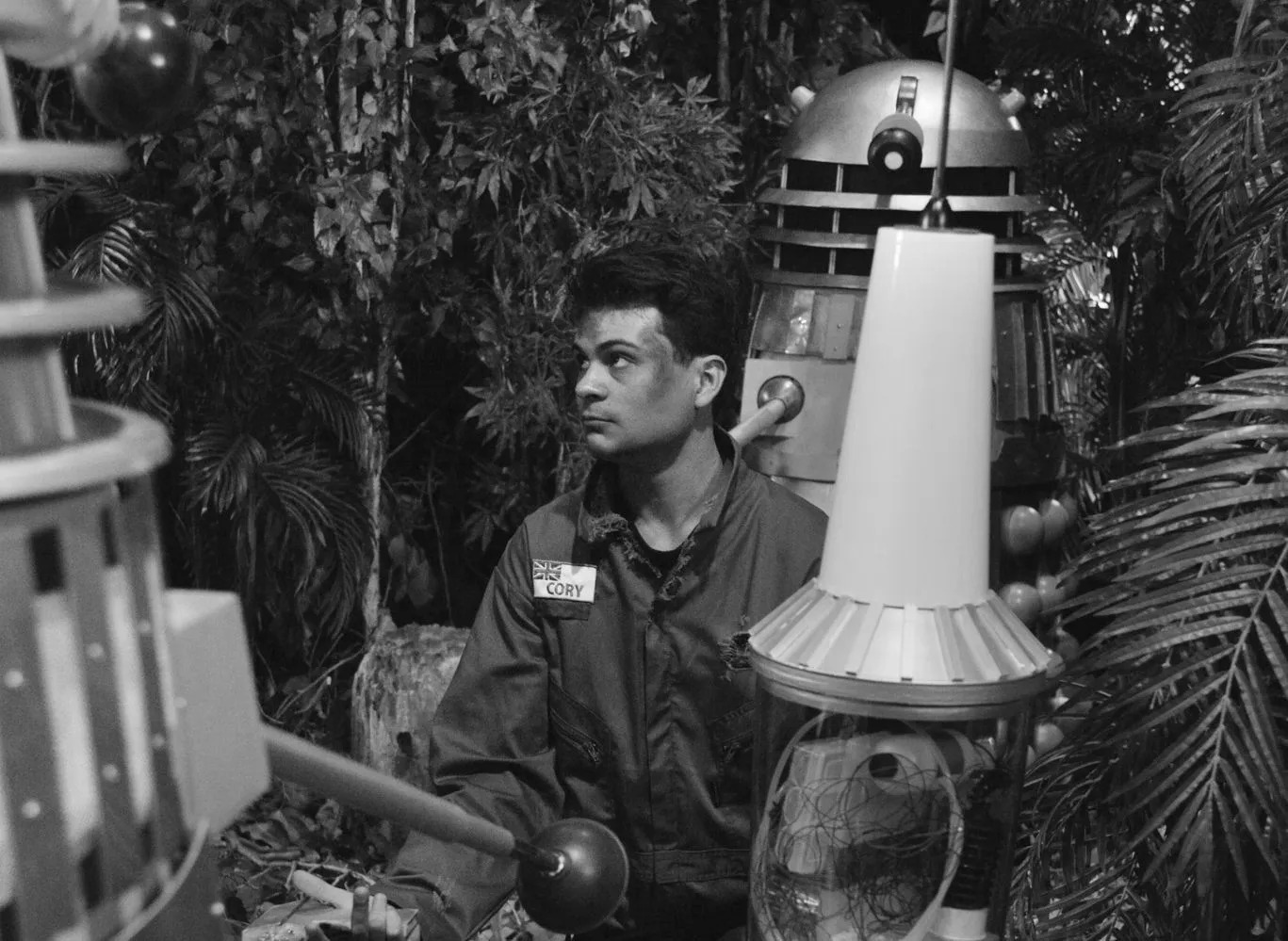 Screencap from Mission to the Unknown (Remake) showing Marco Simioni as Marc Cory, a dashing young man with short dark hair in a jumpsuit. He wears a nametag labeled 'CORY' with a Union Jack. He sits in a forest, looking up at two Daleks around him. Next to him is some sort of small rocket.