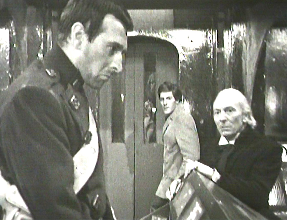Lower-res screencap from Nightmare on Planet Kembel (episode 5: The Traitors): inside a spaceship, Bret Vyon stares down past a control console in profile in the foreground. In the middle ground, the Doctor leans on the same console with a worried expression. Behind him, Steven stands by a futuristic set of doors, looking back. Through one of the door's windows someone can be vaguely seen.