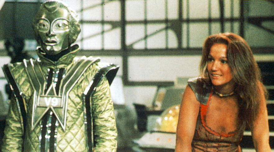 Screencap from The Robots of Death: Leela grins at the camera, leaning over a console, with a robot next to her.