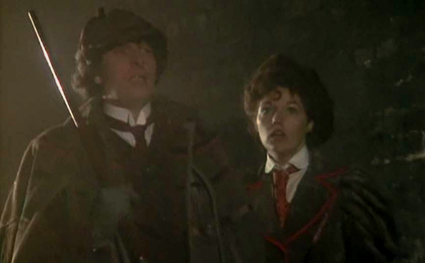 Screencap from The Talons of Weng-Chiang: in a foggy alley, the Doctor (in a deerstalker and tweed coat) and Leela (in a hat and brown red-lined jacket with collar) look past the camera.