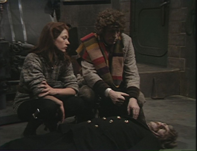 Screencap from Horror of Fang Rock: The Doctor and Leela, in a sweater and pants, crouch beside the body of a man with a mustache in a room with machinery.