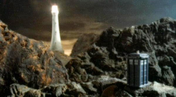 Screencap from Horror of Fang Rock: the TARDIS sits on a rocky plateau. In the background is a lighthouse with its lamp lit.