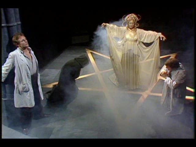Screencap from Image of the Fendahl: in a smoky basement, a golden woman in golden robes stands in the middle of a large pentagram on the floor. Two figures bow to her: one robed, and one in a white coat. A man in a lab coat is tied to a pillar nearby.