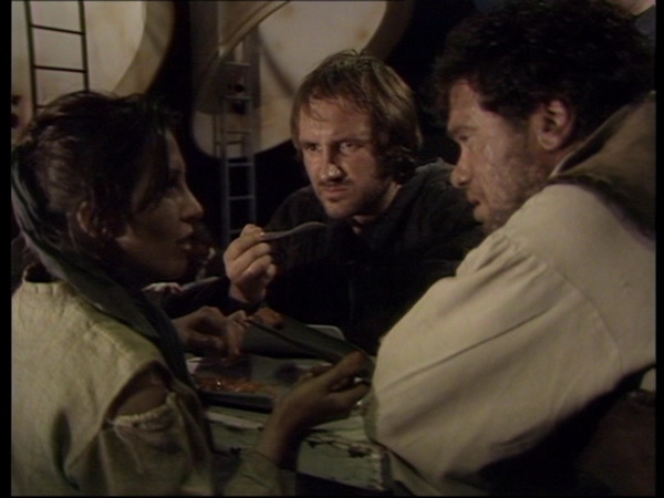 Screencap from The SUn Makers: three people in dirty brown clothes crowd around a small table, eating ambiguous food. The woman nearest to the camera explains something to Mandrel, the man to her right: across from her, another man looks on in distaste.