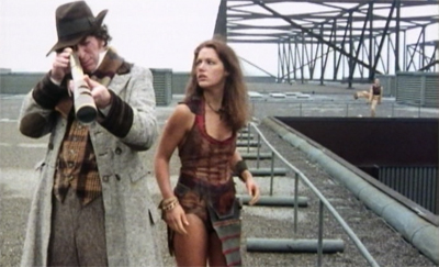Screencap from The SUn Makers: the Doctor and Leela stand on a huge asphalt roof. The Doctor looks down and towards the camera through a huge telescope. In the background, a man in a yellow suit stands near the edge.