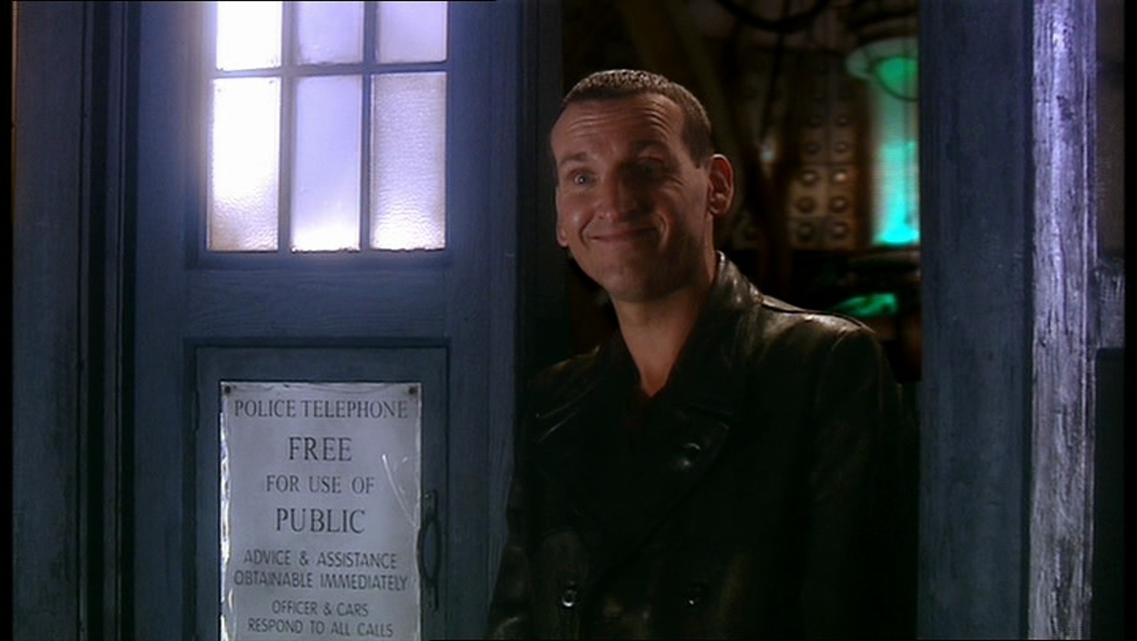 Screencap from Rose: The Doctor (Christopher Eccleston) stands grinning in the doorway of the TARDIS.