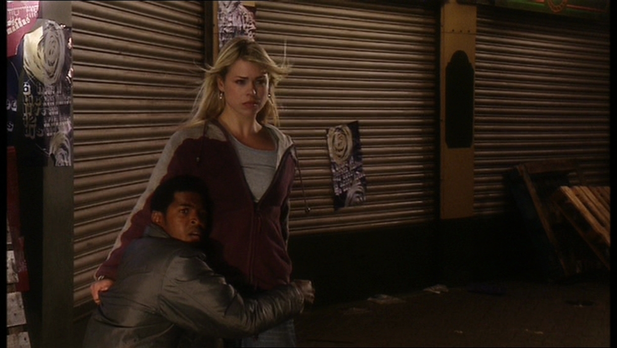 Screencap from Rose: In a back alley of some sort, Rose stares out towards the camera, with Mickey clinging to her legs.