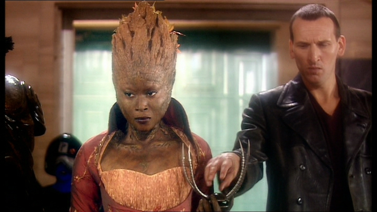 Screencap from The End of the World: Jabe, a tree-woman in a red outfit, holds a mechanical spider upside-down in her hand. The Doctor, standing next to her, reaches for it.
