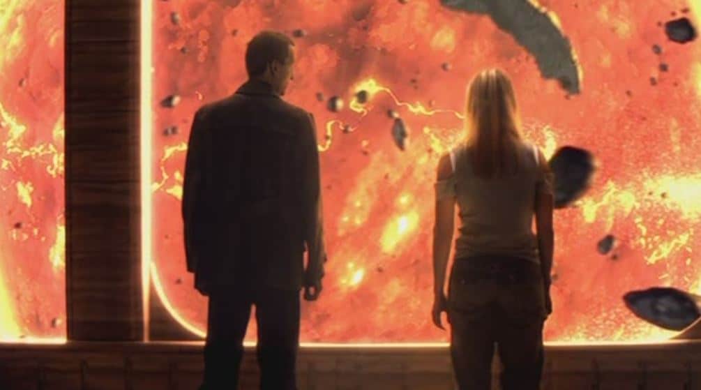 Screencap from The End of the World: the Doctor and Rose stare out a wide floor-to-ceiling window at space. Chunks of debris fly past the window, and a huge, glowing orange sun silhouettes the two, facing away from us.