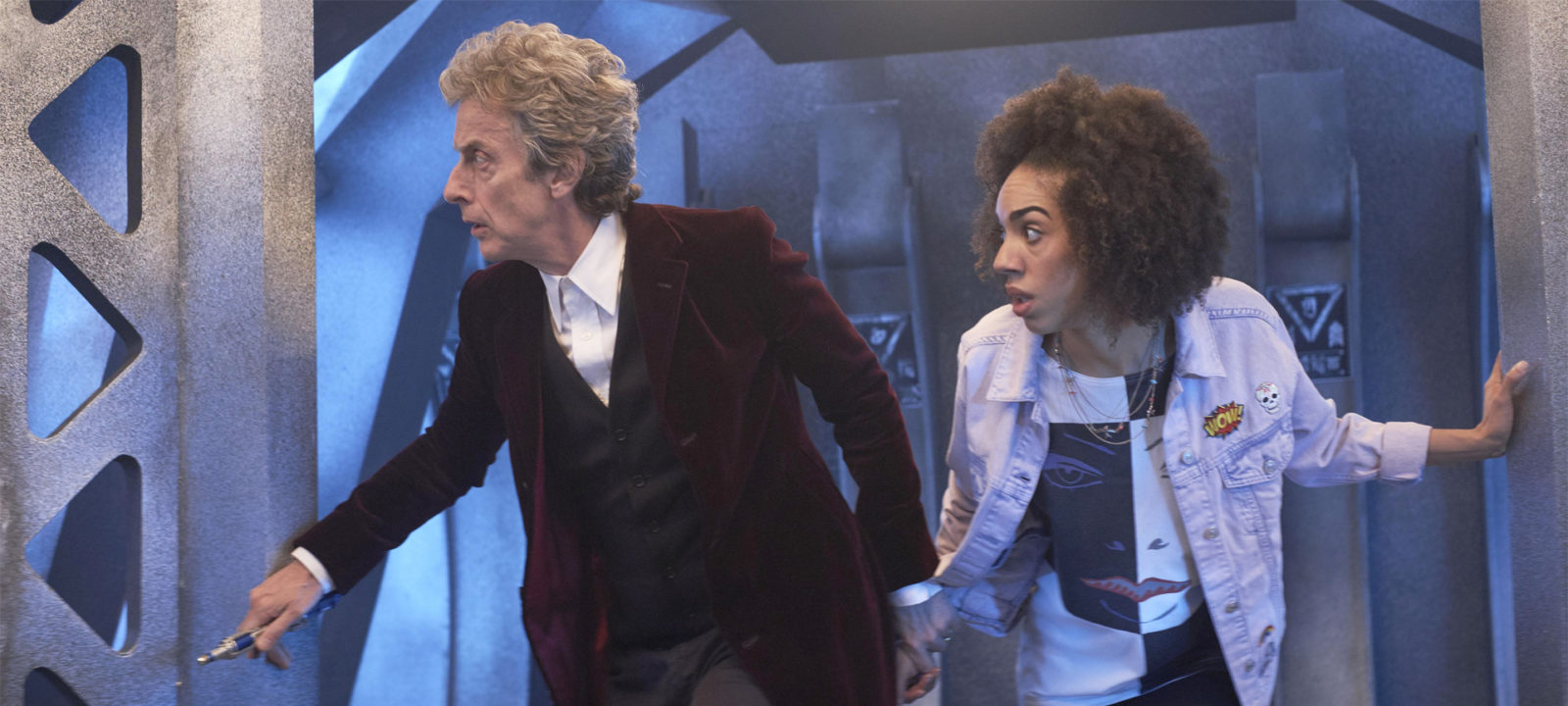 Screencap from The Pilot: the Doctor and Bill in a sort of futuristic doorway, next to a girder. Both look off to the left of the screen, and the Doctor holds Bill's wrist.