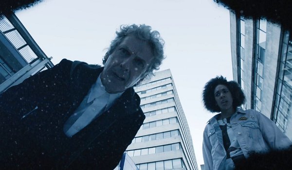 Screencap from The Pilot: the reflections of the Doctor and Bill looking at a puddle on some pavement. Several tall modern buildings are in the background. Bill is a young black woman with an afro in a lavender jean jacket with a few patches.