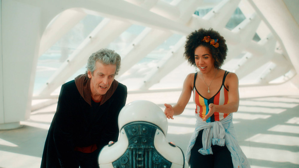 Screencap from Smile: the Doctor and Bill stand in a bright white futuristic city, crouching down to look at a short robot that faces away from the camera.