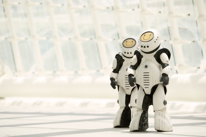 Screencap from Smile: two robots stand in a futuristic white city. The robots are shorter than humans and chunky in design, black with white panelling covering most of their bodies. Their faces are yellow screens which display simplistic smiley faces. The eyes in the faces are exclamation marks.