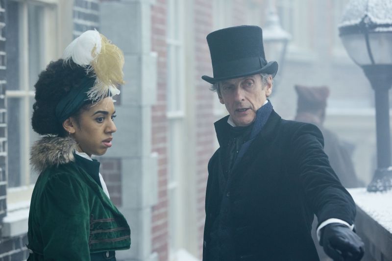 Screencap from Thin Ice: the Doctor and Bill stand in a snowy street in Victorian London. The Doctor wears a black suit with top hat, and Bill wears a dress with a green velvet jacket and a tall feathered hatpiece.
