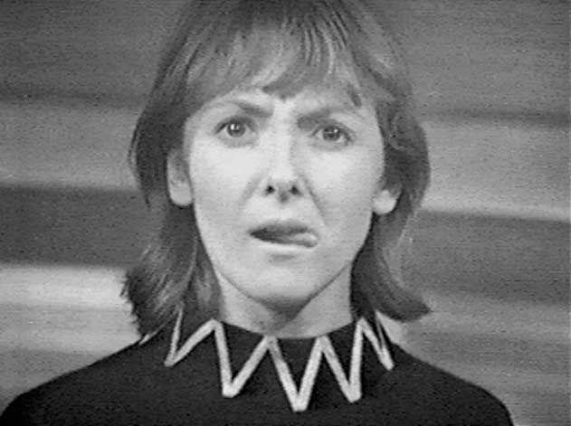 Screencap, possibly from The Rescue: Vicki stares at a point above the camera with a calculating or thoughtful look, tongue poking out the side of her mouth. She wears a black sort of smock with a high collar which has a zigzag pattern on it, and her hair is down, resting around her shoulders.